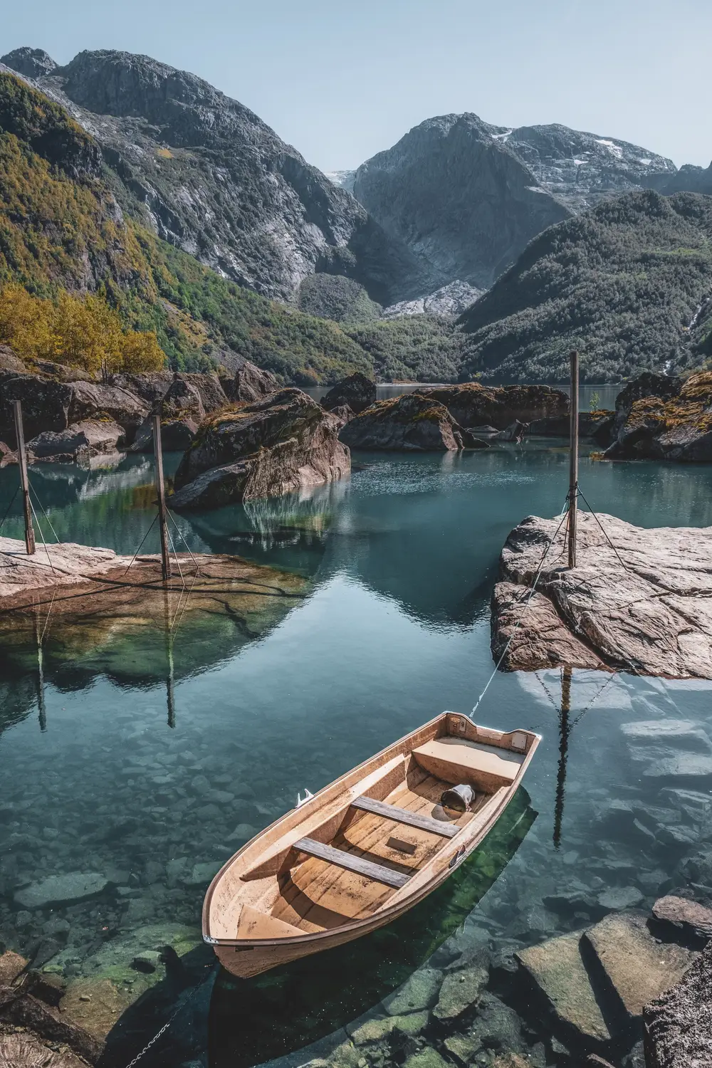 A wooden rowboat floating on crystal-clear turquoise water, with rugged mountains rising behind the rocky shore.