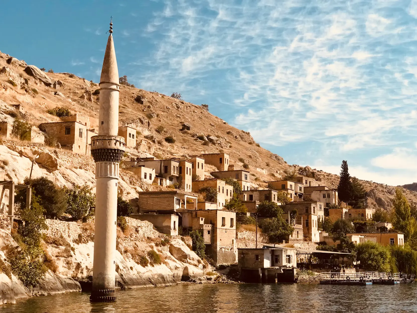 A tall minaret and hillside village of stone and adobe buildings overlooking a river, with a partly cloudy sky in the background.