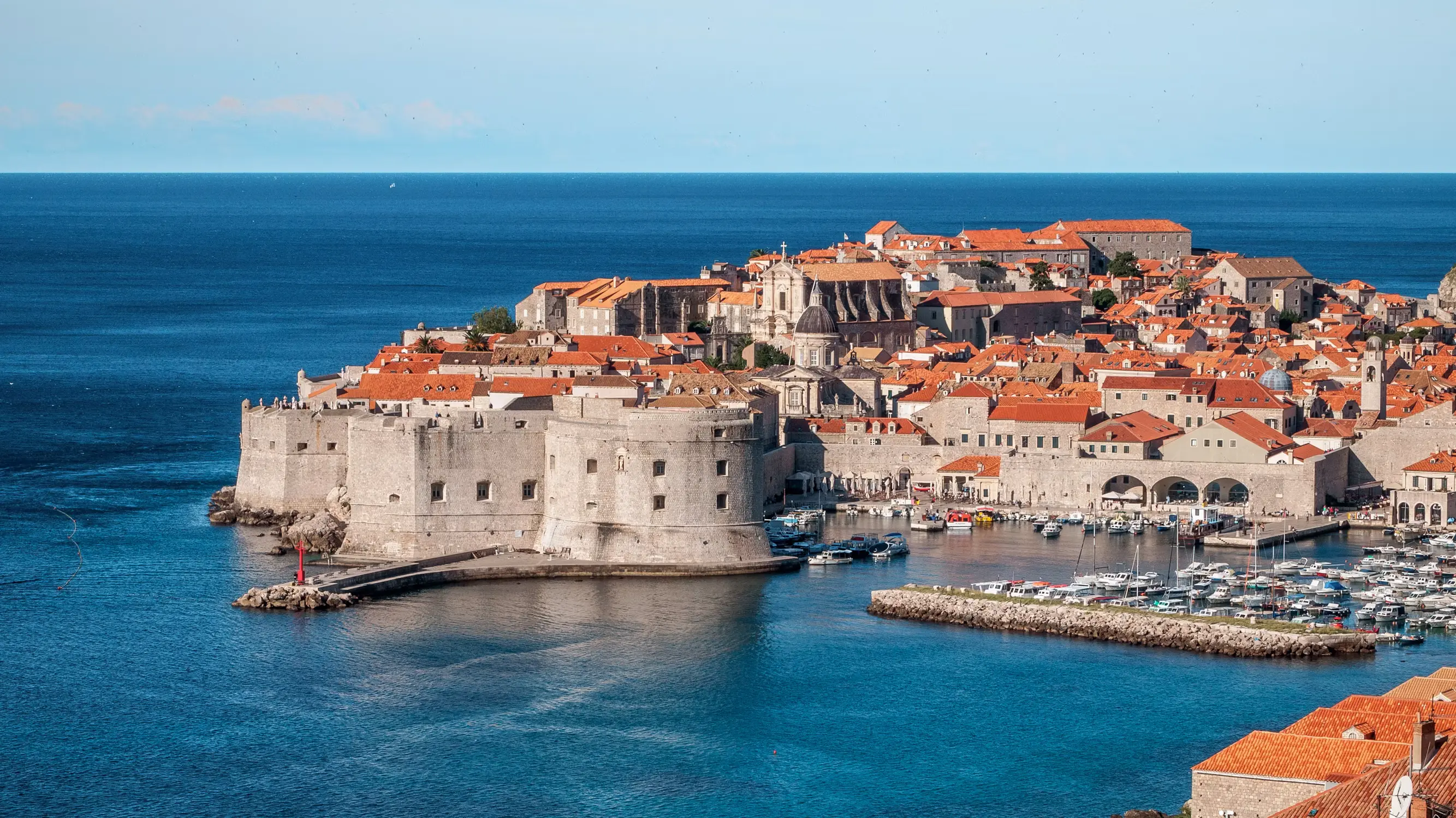 Aerial view of Dubrovnik's old town peninsula with red-roofed buildings, city walls, and a marina, surrounded by deep blue sea.