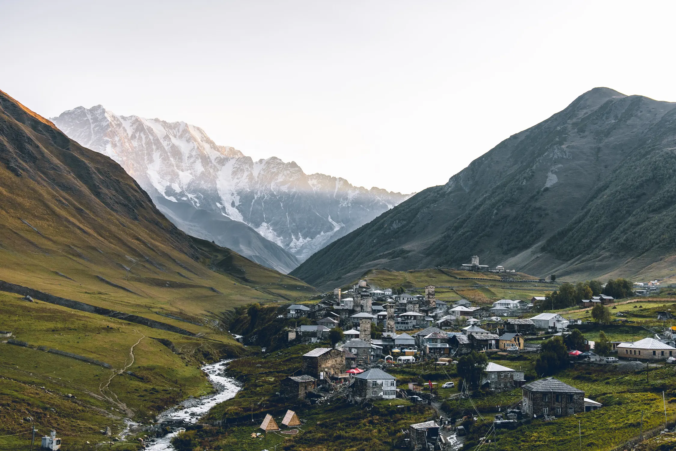 Mountain village in a green valley with a winding stream and snow-capped peaks in the background.