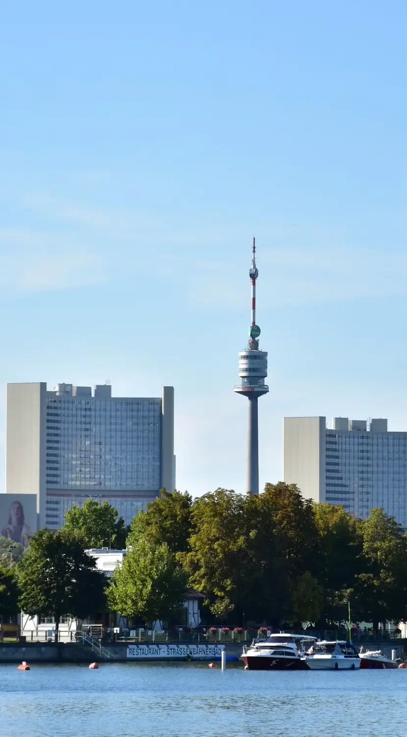 A city skyline viewed across a river, featuring a tall slender tower and modern high-rise buildings under a clear sky.