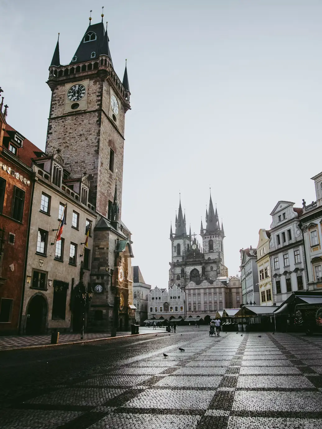 Old Town Square at dusk with the clock tower and Týn Church spires along an empty checkered street