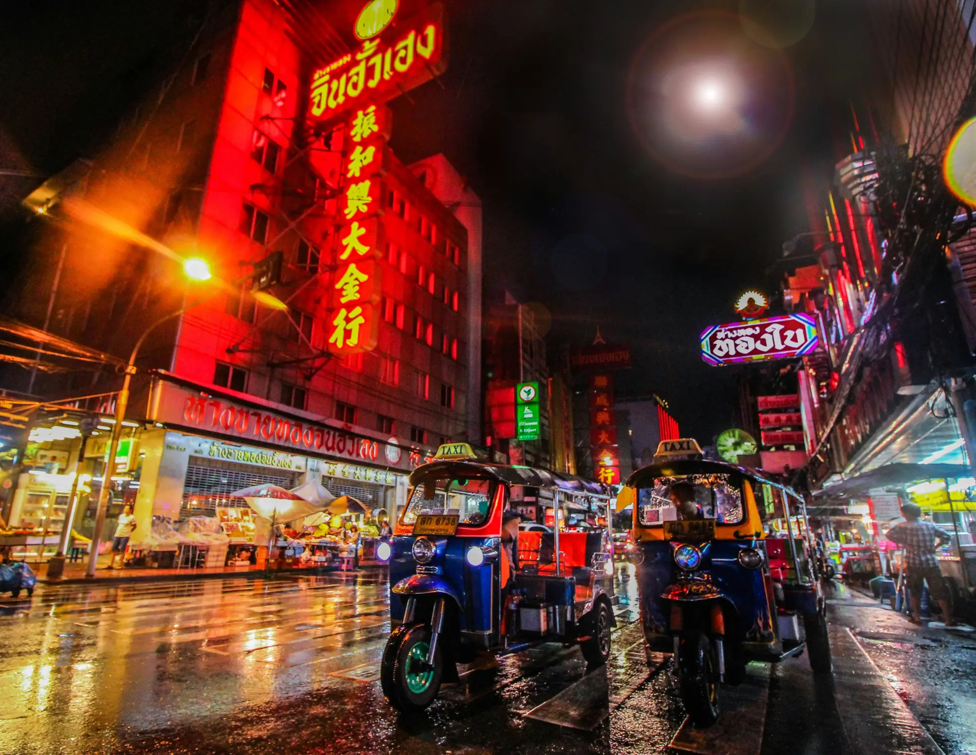 Busy Bangkok Chinatown street at night, with colorful neon signs, food stalls, wet reflective pavement, and tuk-tuks parked along the road.