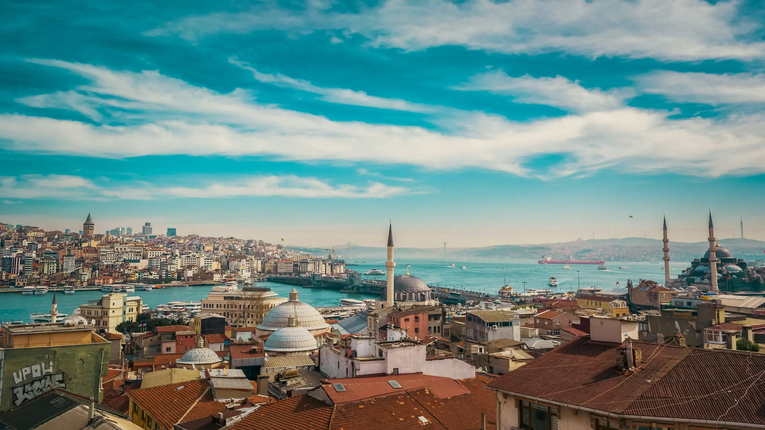 An elevated view of Istanbul's rooftops and the Golden Horn, with a domed mosque and minarets nestled among the buildings under a bright blue sky.