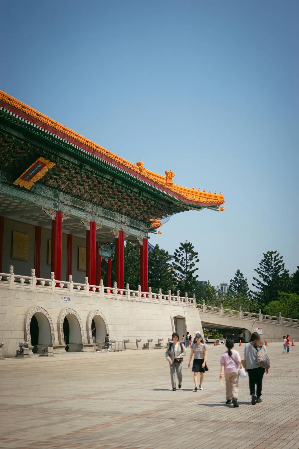 People walking past the National Concert Hall in Taipei, with its golden tiled roof and red columns under a clear blue sky