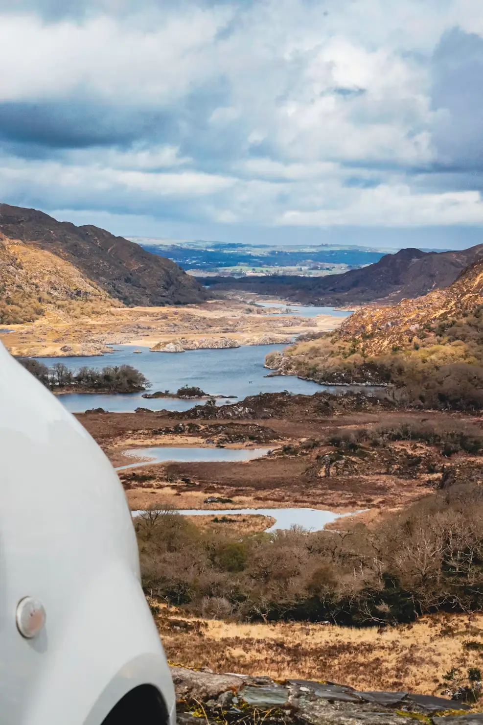 Mountain lake landscape with rolling hills and clouds, viewed from inside a vehicle, Ireland.