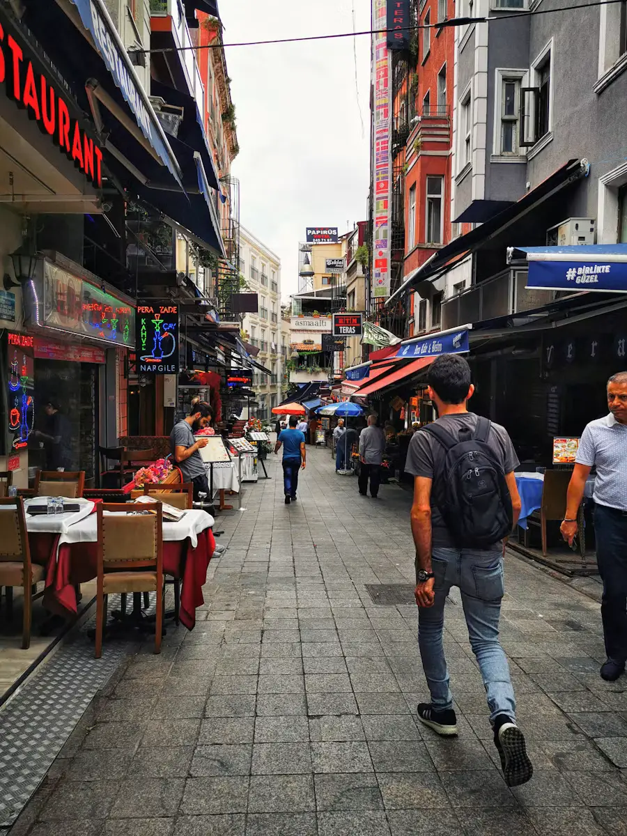 A pedestrian street lined with restaurants and outdoor seating, with people walking past shops and buildings.