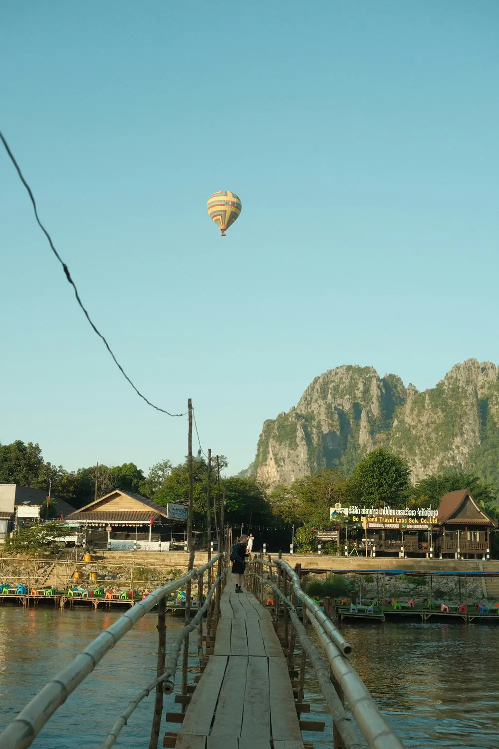 Hot air balloon floating above karst mountains and a river, viewed from a wooden bamboo footbridge in Vang Vieng, Laos.