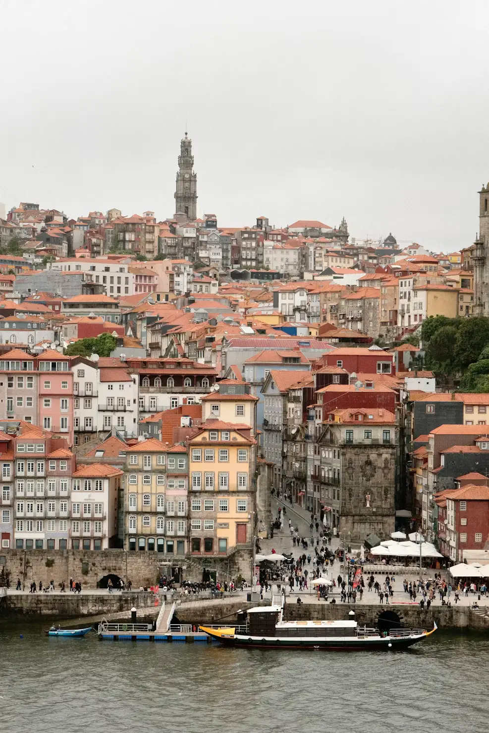 Historic riverside city with colorful buildings, terracotta rooftops, a tall bell tower, and boats along the waterfront.