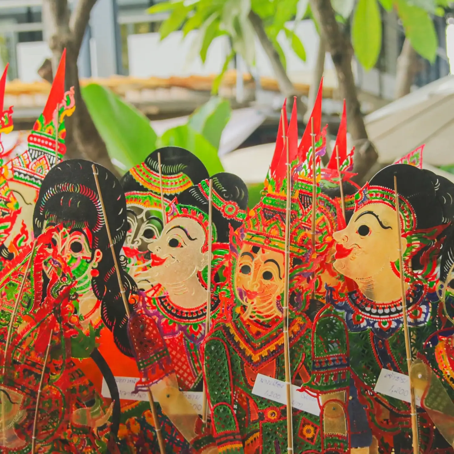 Colorful traditional shadow puppets with ornate headdresses displayed on sticks against a leafy backdrop.