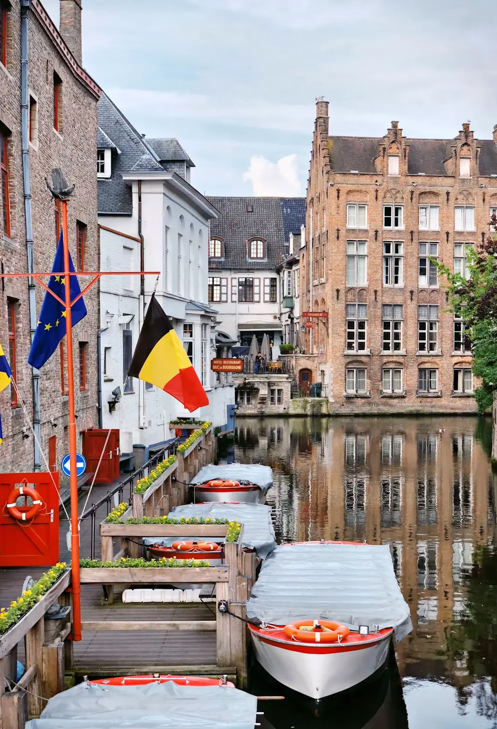 Tour boats moored along a Bruges canal lined with brick buildings, with Belgian and EU flags flying from a red pole.