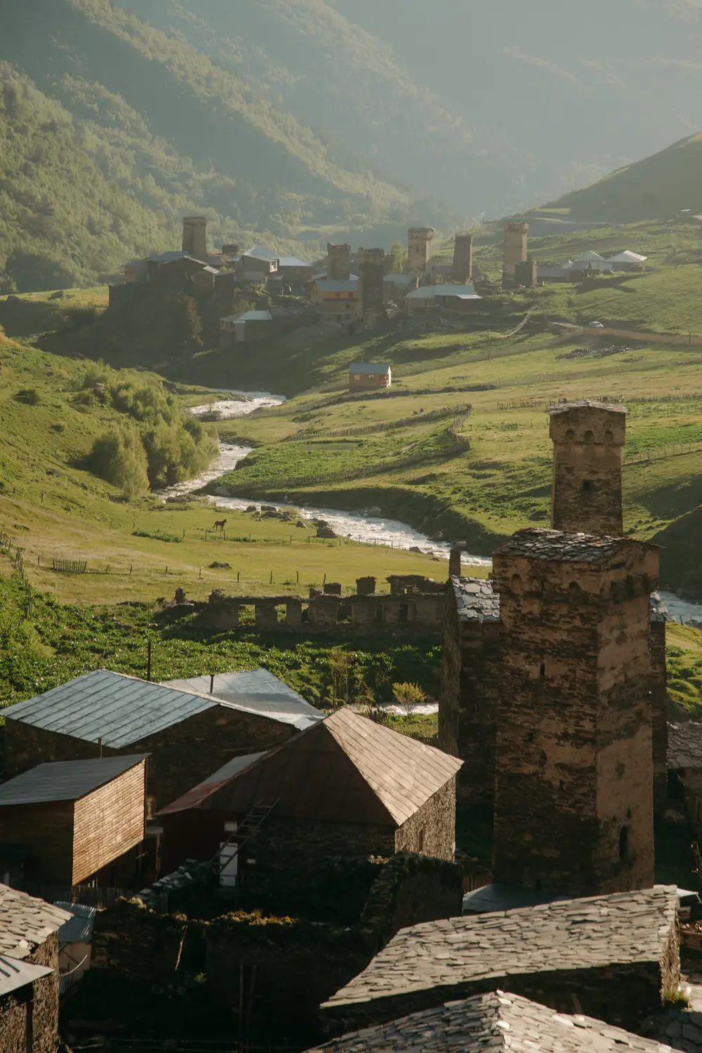 A traditional mountain village with stone towers and slate-roofed houses, surrounded by green fields and forested mountains, with a winding river through the valley.