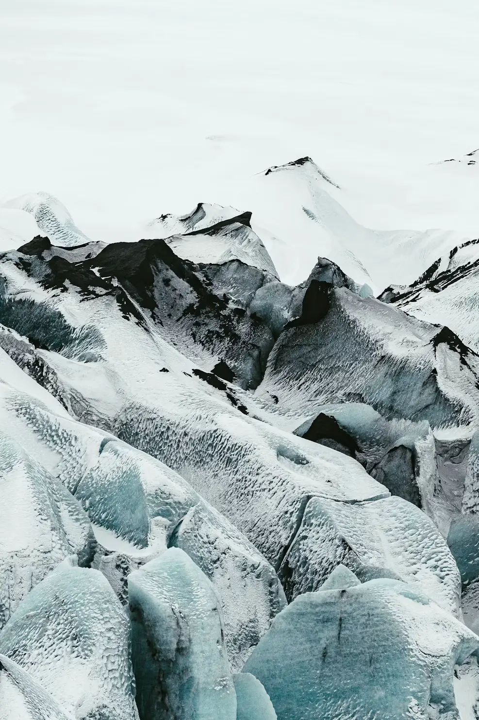 Rugged glacier landscape with textured blue-white ice and dark crevasses beneath snow-capped peaks, Iceland.