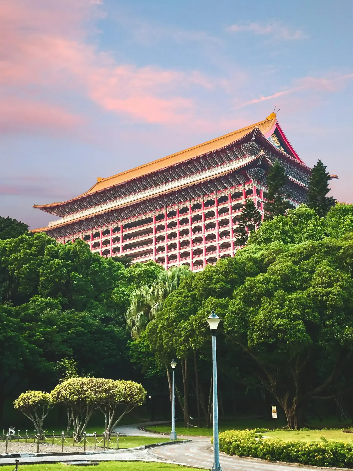 The Grand Hotel in Taipei, a red palatial building with a golden tiled roof, rising above green trees under a pink sunset sky