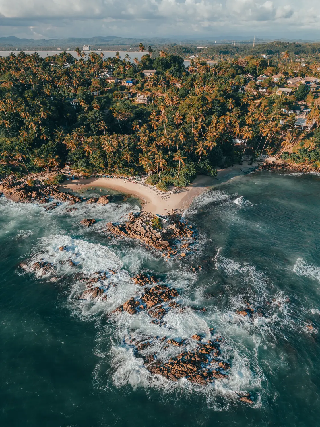 Aerial view of a rocky palm-fringed coastline with waves crashing against the shore in Sri Lanka.