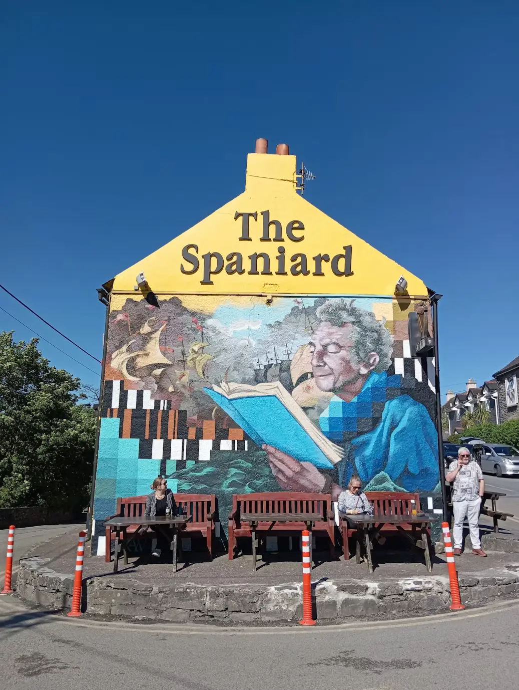 Colorful mural of a couple painted on the yellow gabled facade of a pub called 'The Spaniard', with outdoor seating below.