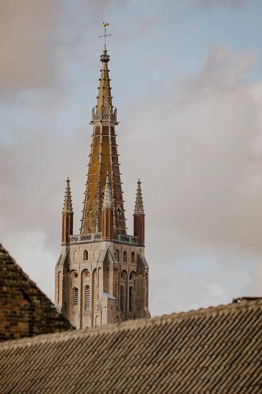 Close-up of the Church of Our Lady's brick spire in Bruges, rising above a tiled rooftop against a cloudy sky.
