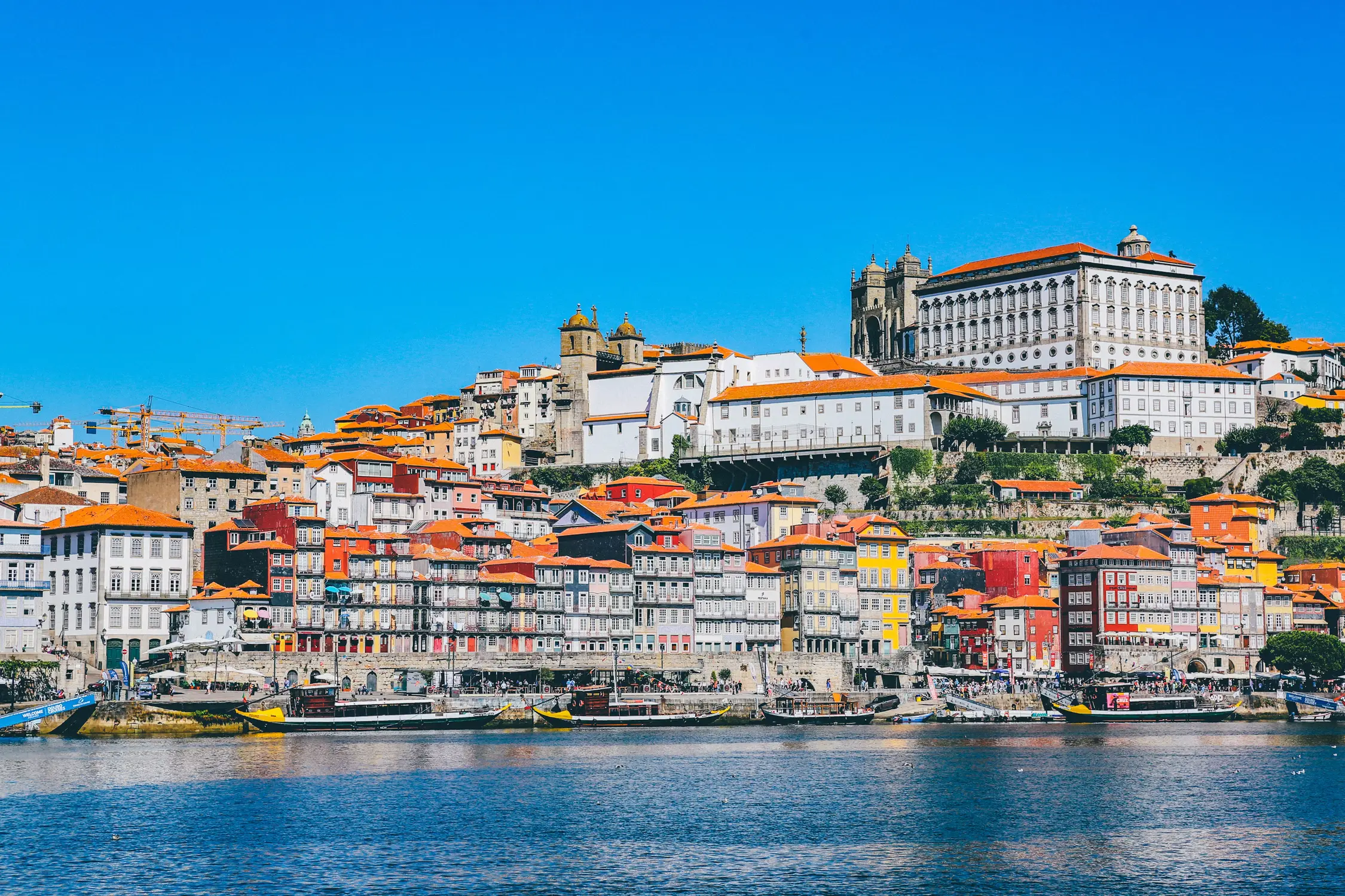 Colorful riverside cityscape with orange-roofed buildings, historic churches, and waterfront boats under a clear blue sky.