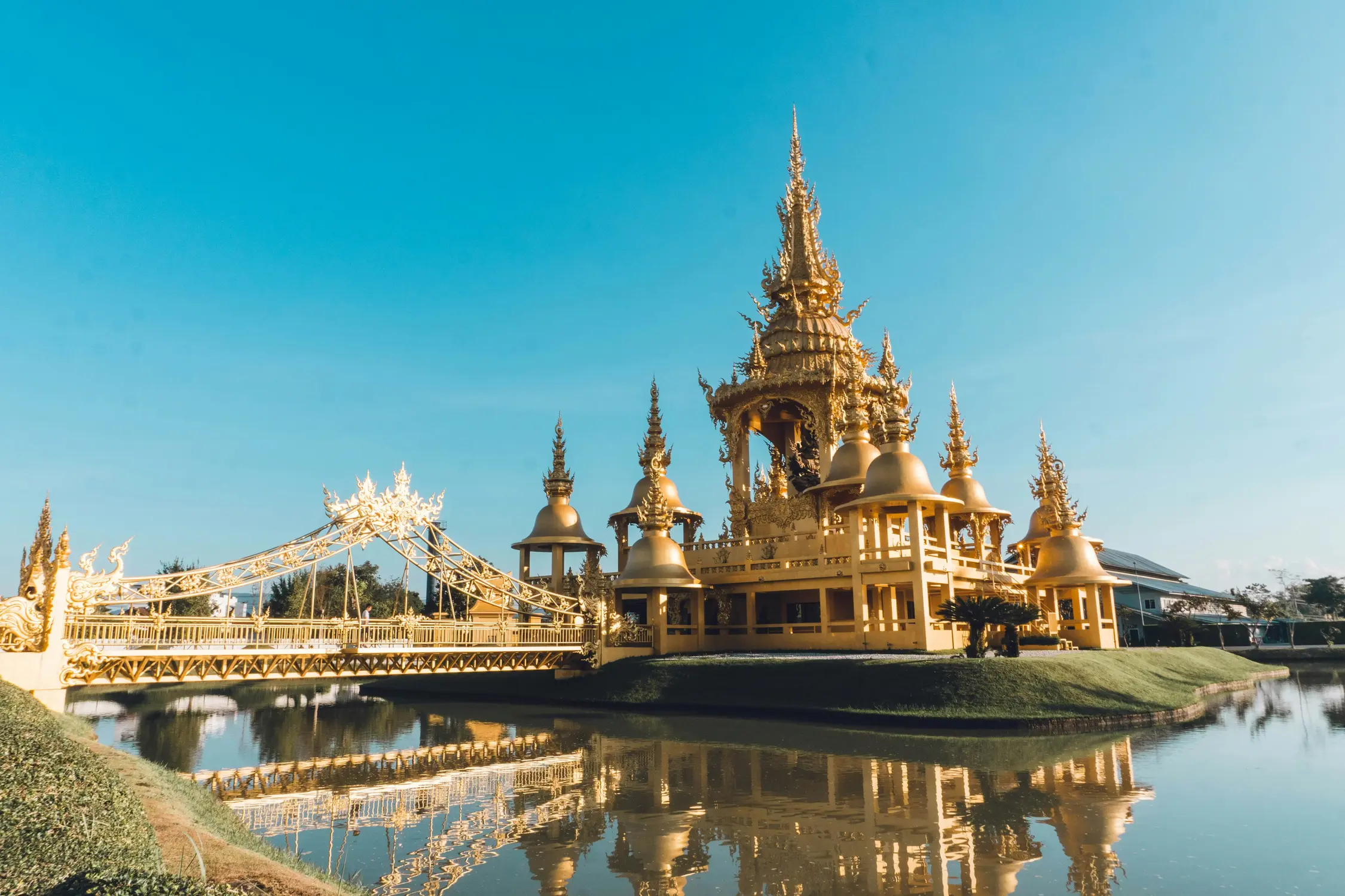 Golden Buddhist temple complex with ornate spires reflecting in a still water pool under a clear blue sky, Thailand