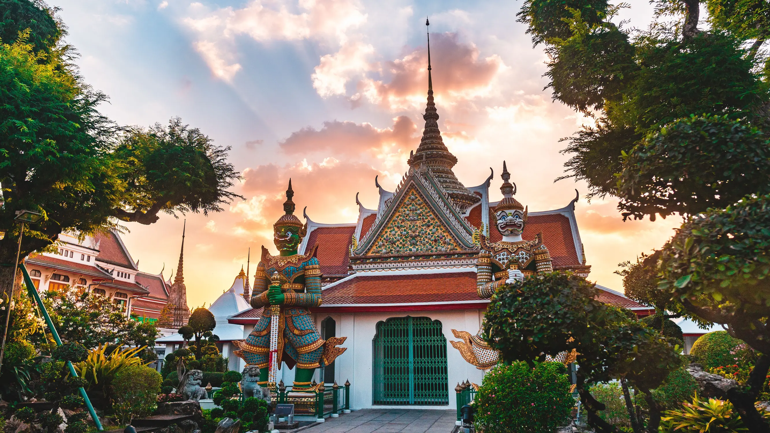 A richly decorated Thai temple at sunset, featuring ornate red-and-gold roofs, intricate mosaic details, towering guardian statues at the entrance, manicured tropical gardens, and a dramatic sky illuminated by warm golden and pink light.