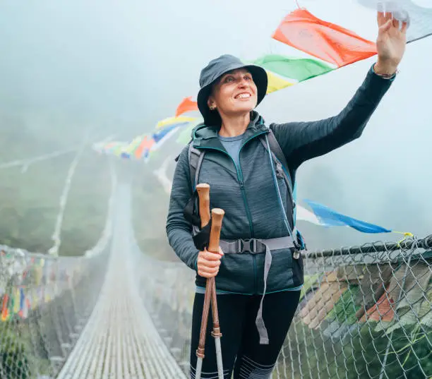 Smiling trekker on a suspension bridge reaching up to colorful Buddhist prayer flags, Nepal