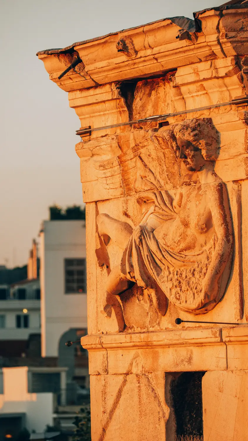 A weathered stone relief carving of a robed figure set into an archway, framed by colorful Greek island buildings.