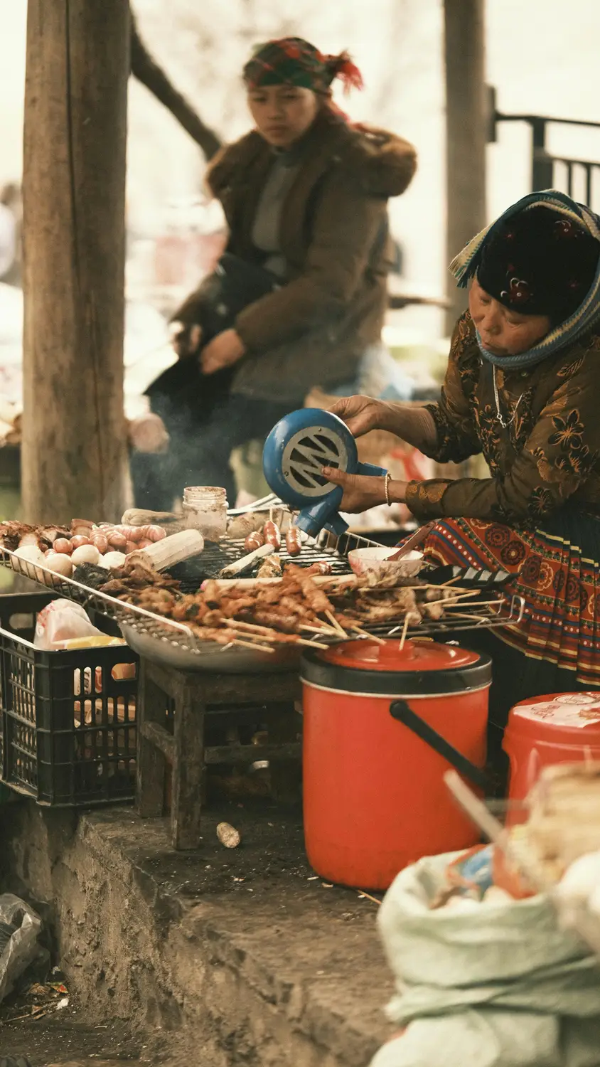 A woman in traditional embroidered dress grilling meat skewers and eggs on a smoking charcoal grill at a busy outdoor street market