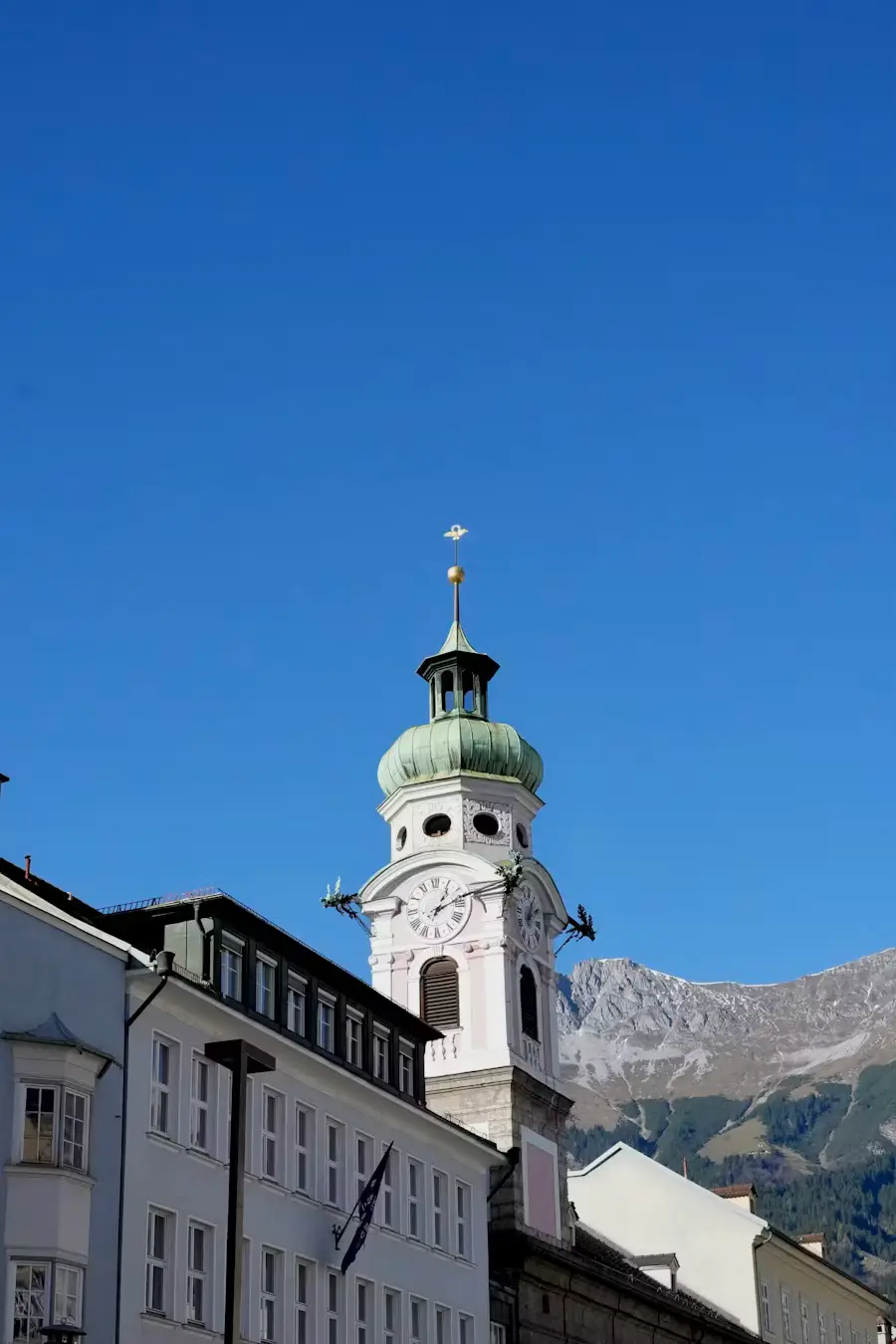 A church tower with a green onion dome rising above rooftops, set against a backdrop of snow-capped mountains and clear blue sky.