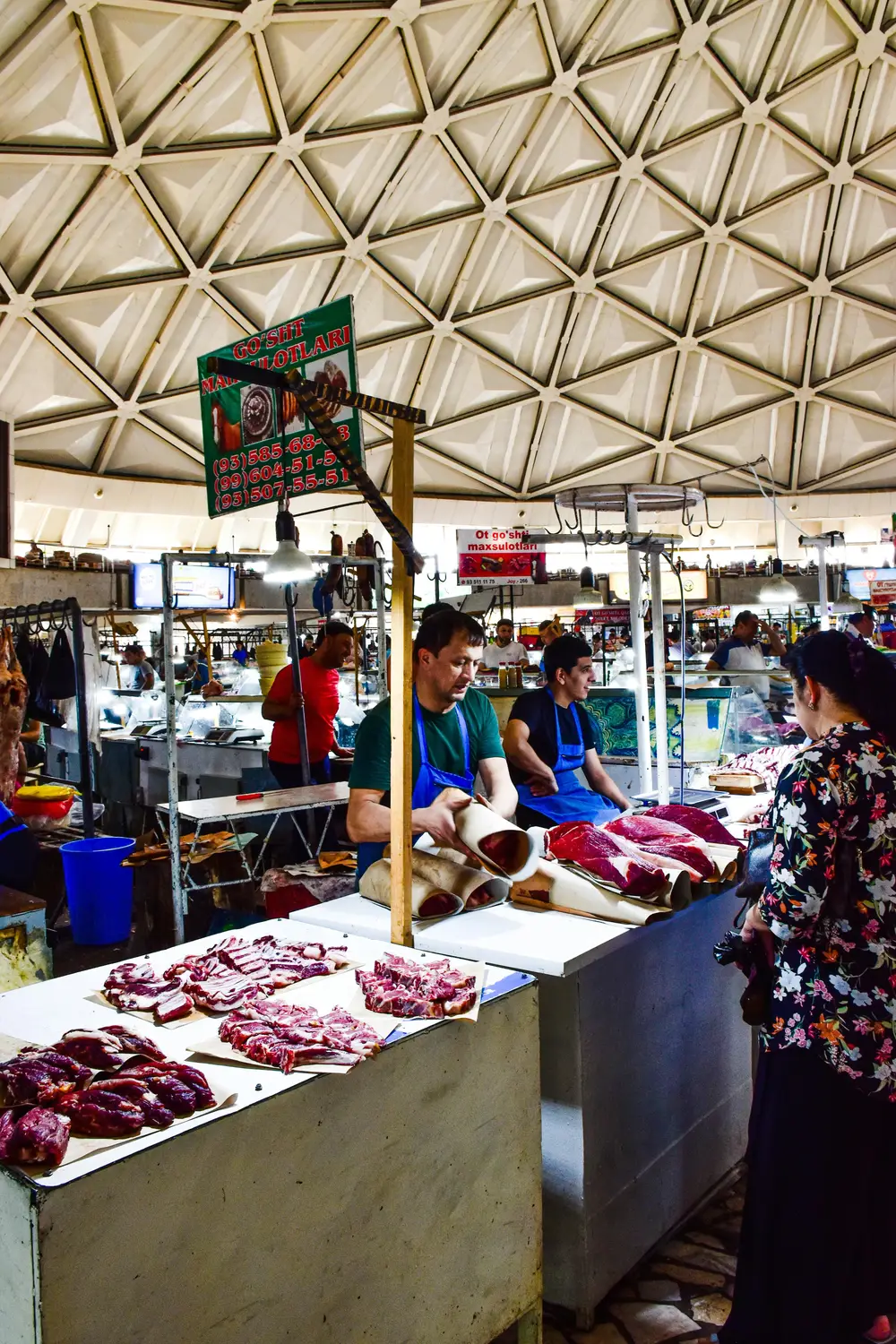 Vendors in blue aprons serving fresh cuts of red meat at a bustling indoor bazaar beneath a striking geometric white dome ceiling