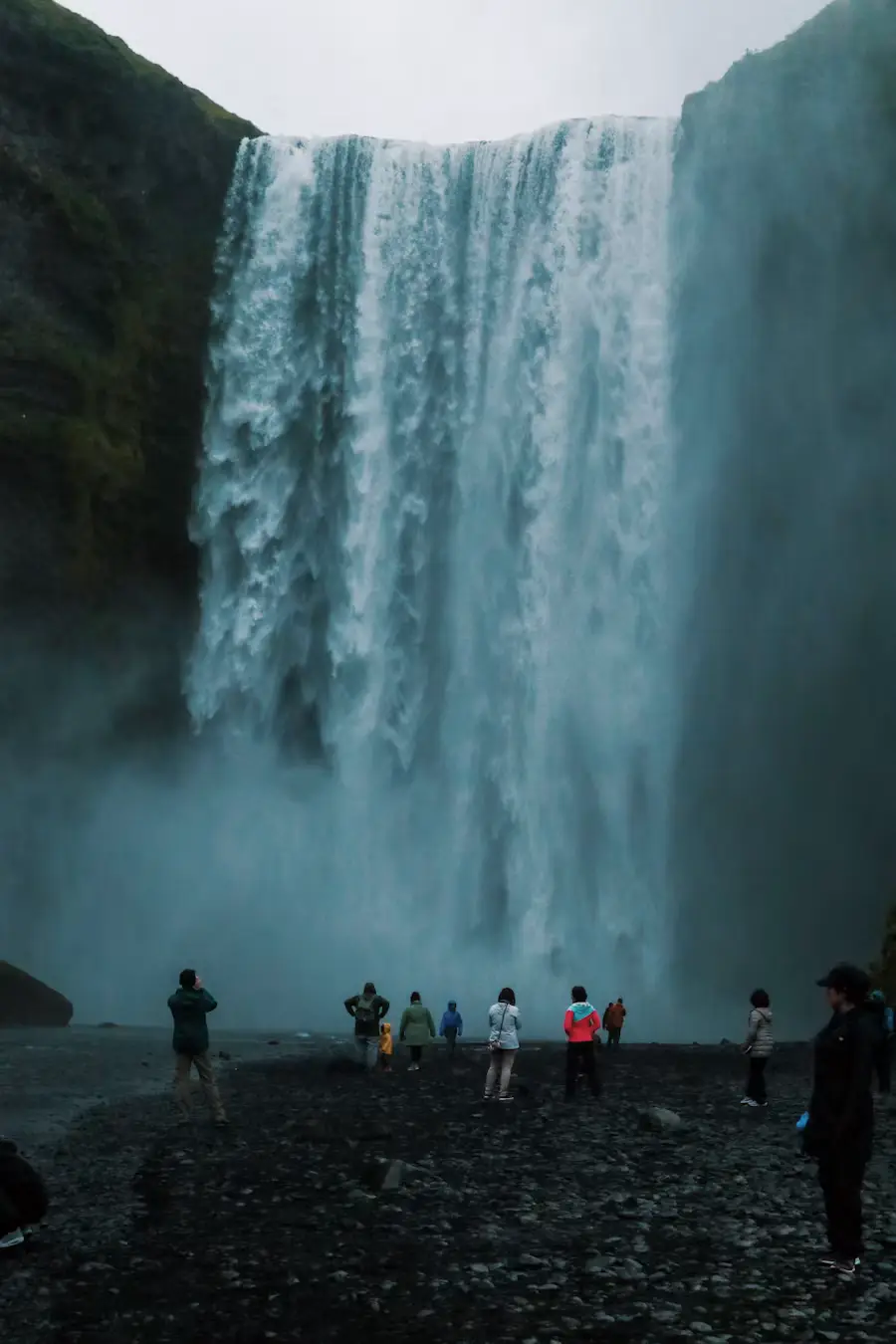 People standing at the base of a powerful waterfall on a rocky beach in dim light, Iceland.