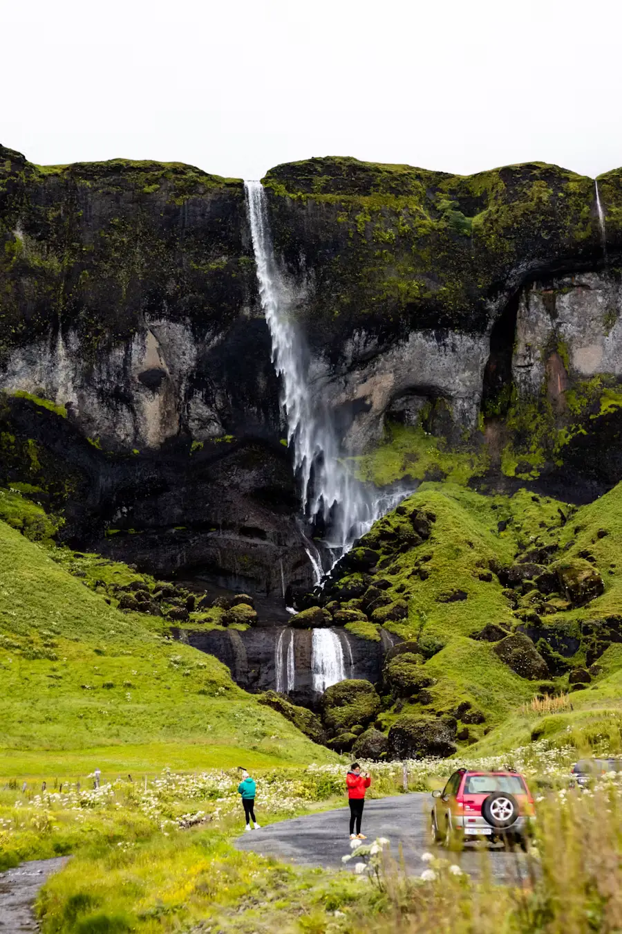 Waterfall pouring from a moss-covered cliff into a rocky gorge, with a car and people visible below, Iceland.