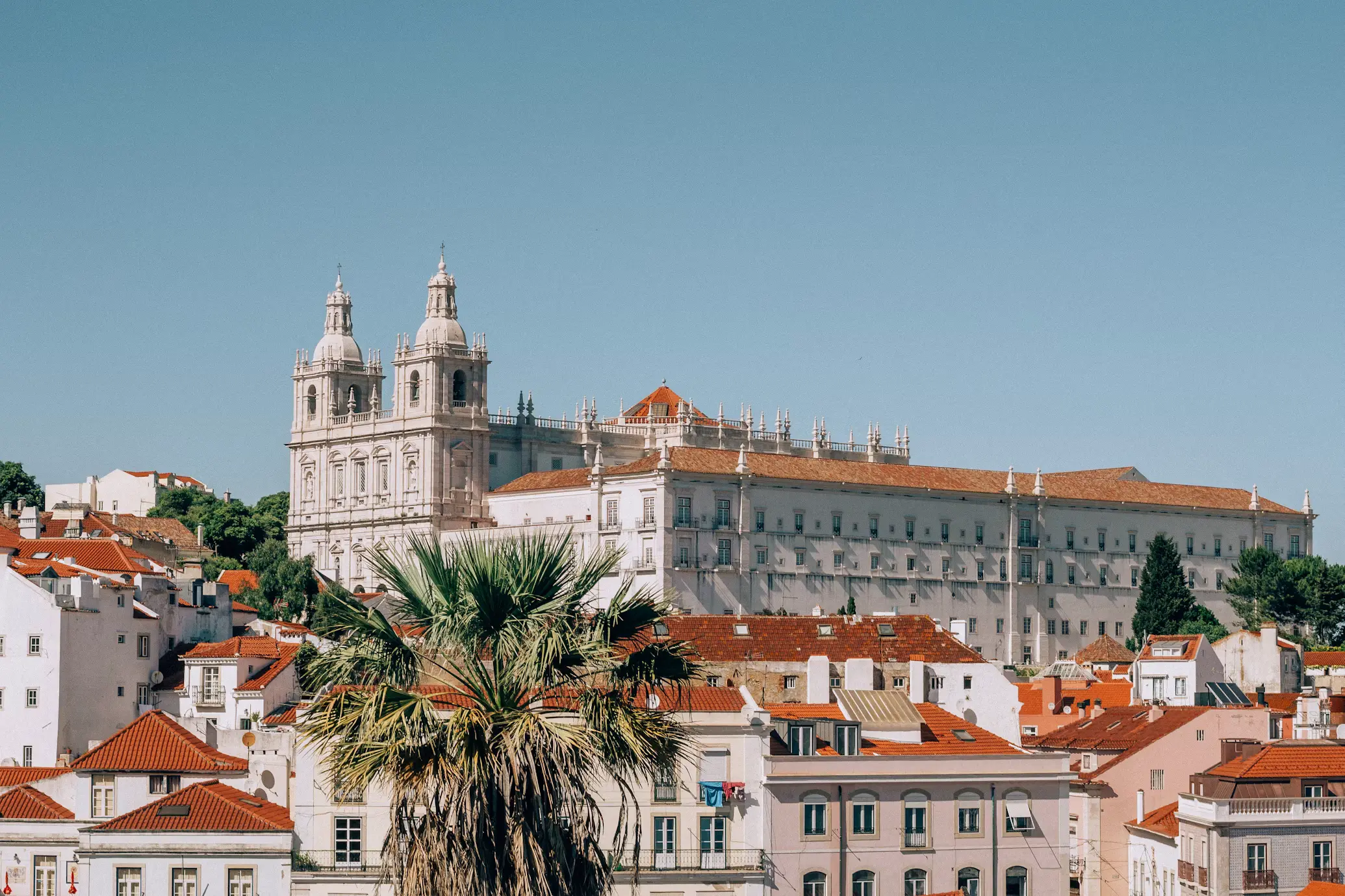 Large white monastery overlooking a city of red-tiled rooftops and palm trees under a clear blue sky.