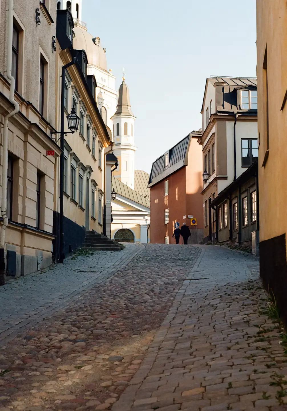Narrow cobblestone alley between old European buildings, with a church spire visible in the distance.
