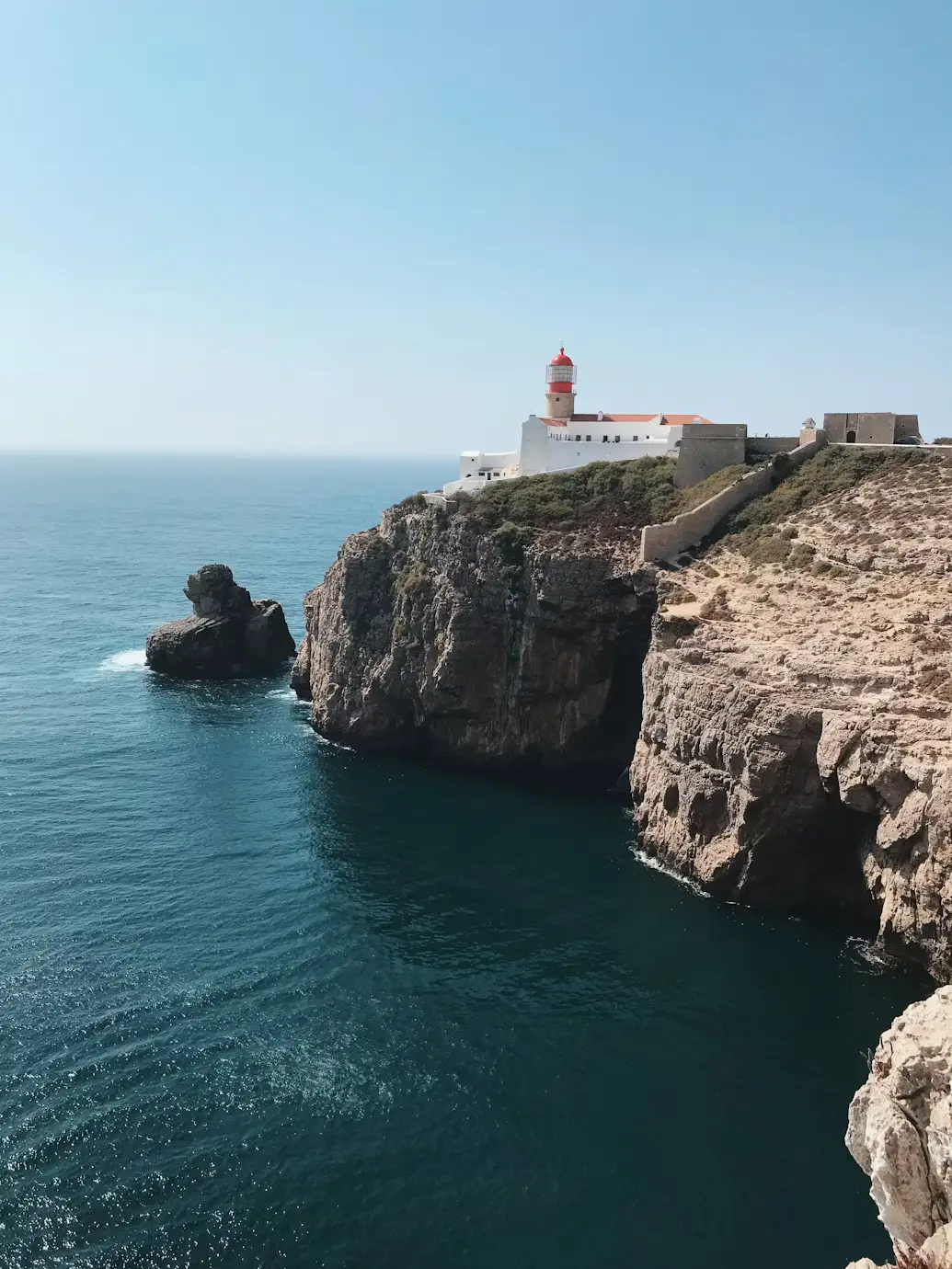Lighthouse atop a white fort on rugged coastal cliffs overlooking the deep blue ocean.