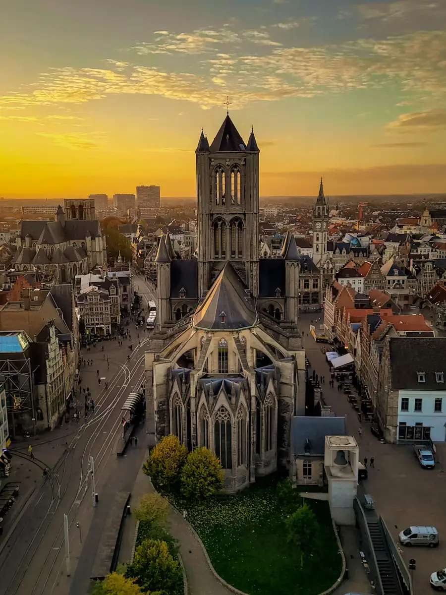 Aerial sunset view of Saint Nicholas' Church tower rising above Ghent's old town rooftops, with a golden sky and the Belfry of Ghent visible in the background.