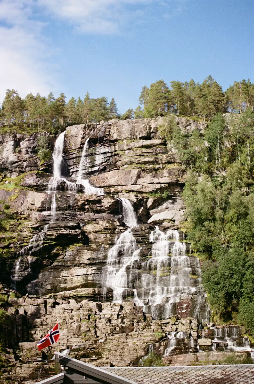 A waterfall cascading down a rocky cliffside surrounded by greenery, with a small Norwegian flag in the foreground.