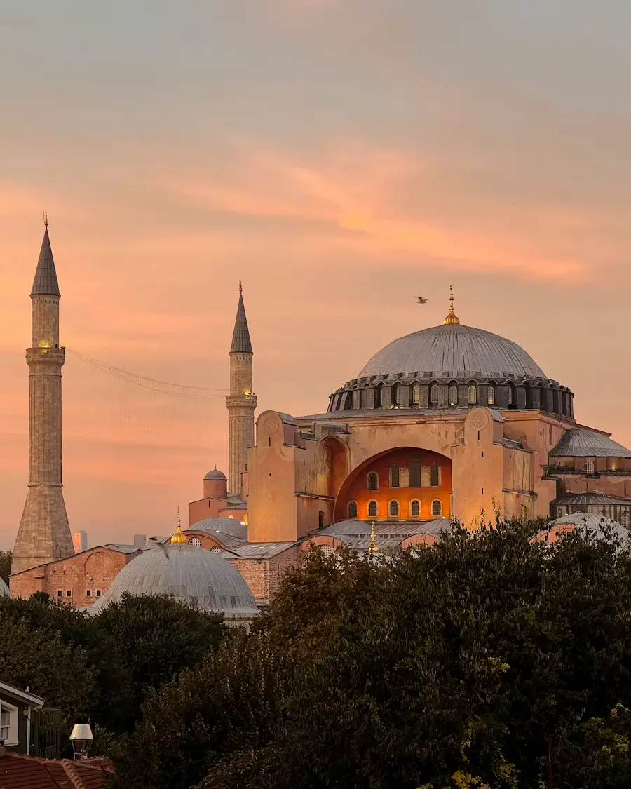 Hagia Sophia's domes and minarets silhouetted against a warm sunset sky, framed by trees in the foreground.