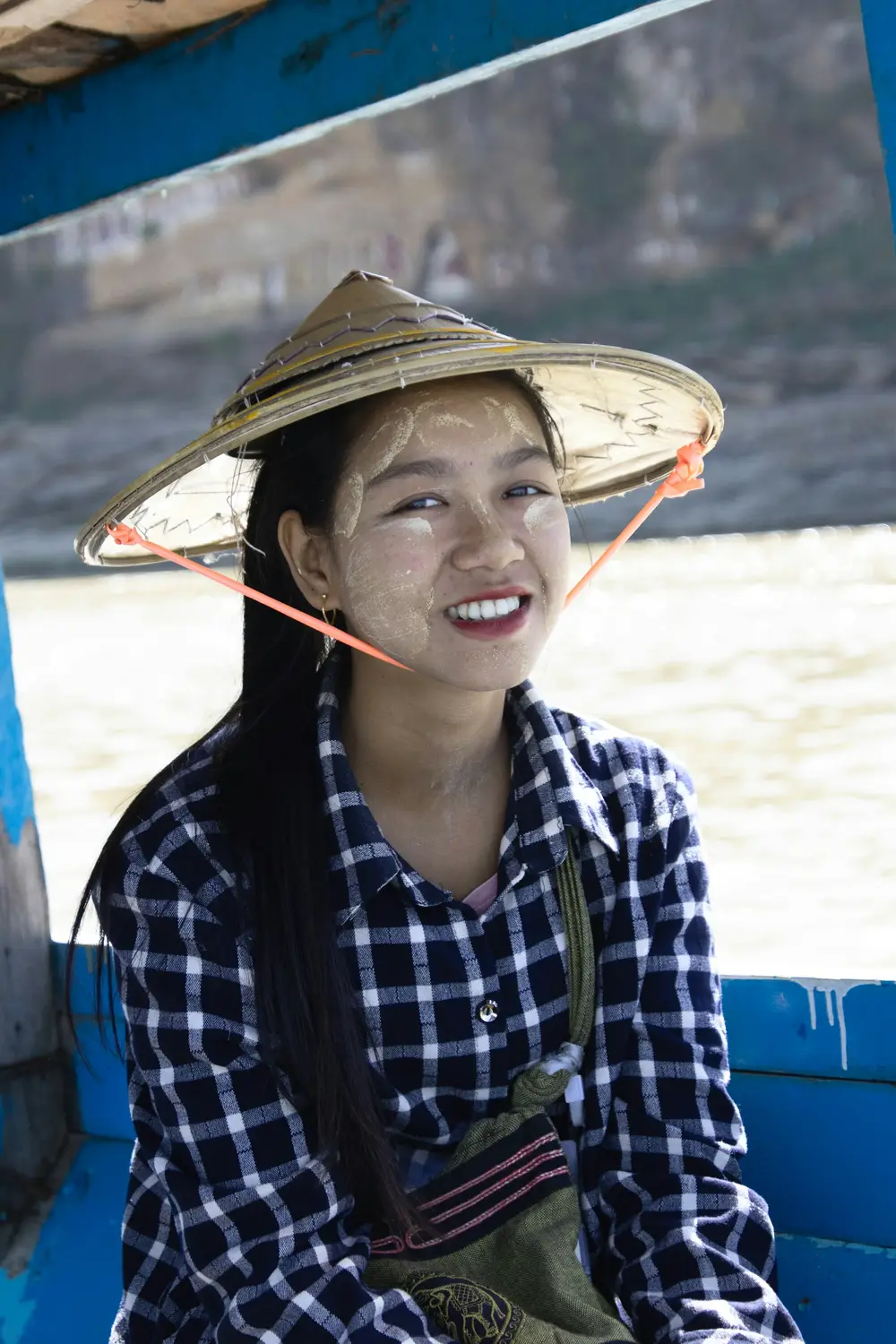 Smiling young Burmese woman wearing traditional thanaka face paste and bamboo conical hat on river boat, Myanmar
