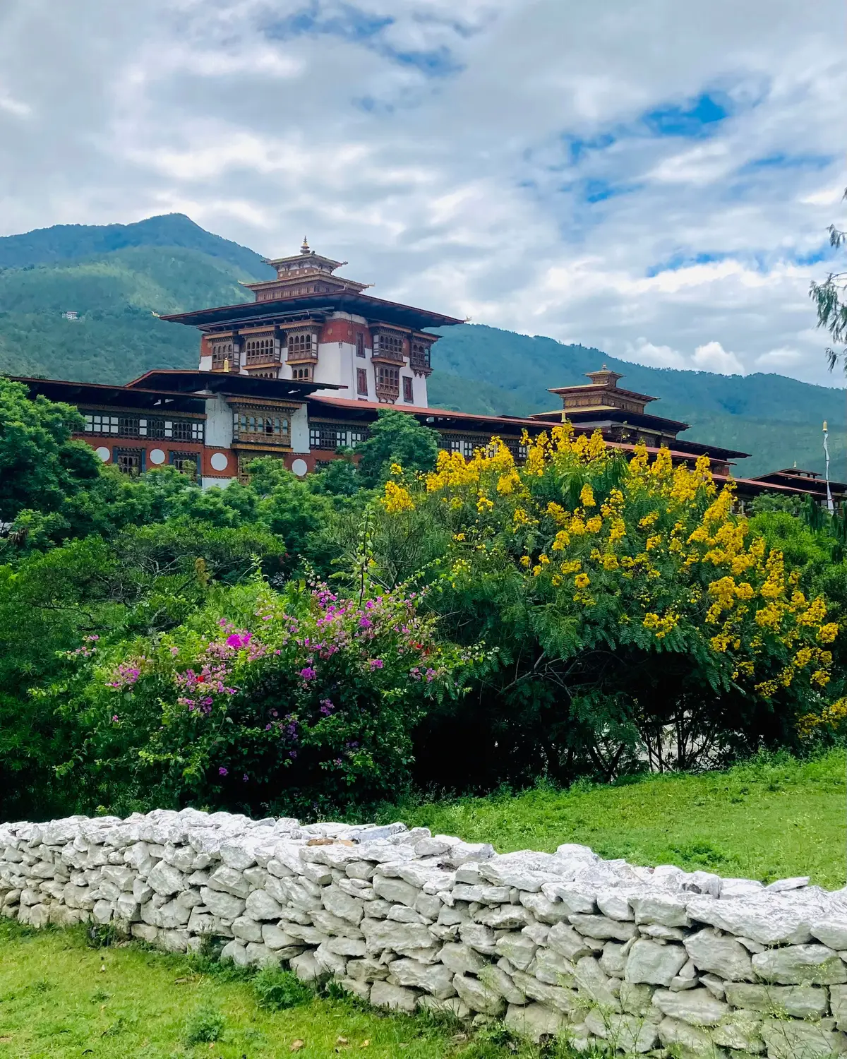 Traditional Bhutanese dzong set on a green hillside with flowering trees and a stone wall against mountains in Bhutan.