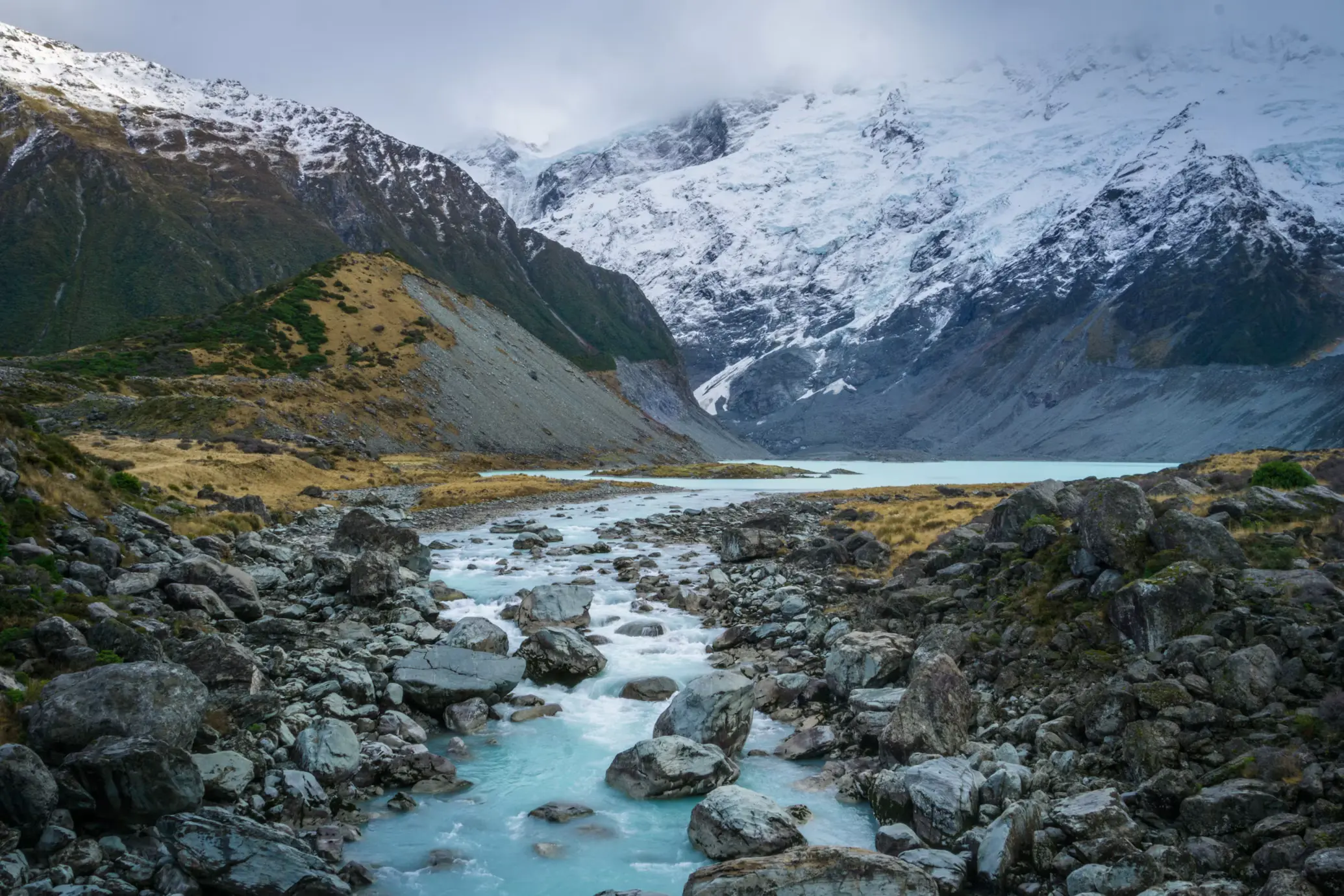 Milky blue glacial river flowing through a rocky valley toward snow-covered mountains beneath low clouds.