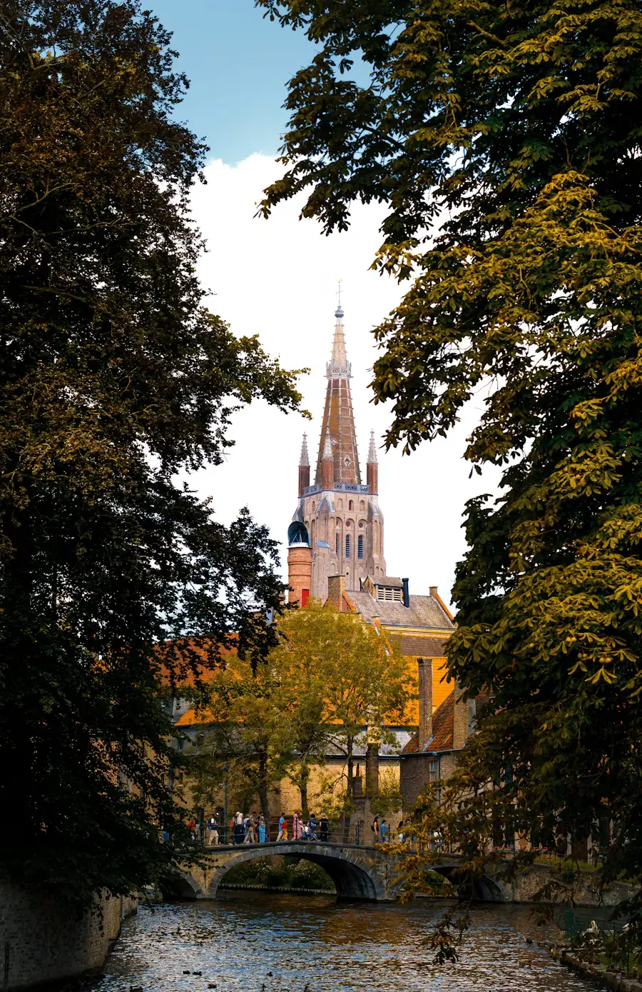 The spire of the Church of Our Lady in Bruges glimpsed through autumn trees above a stone bridge over the canal.