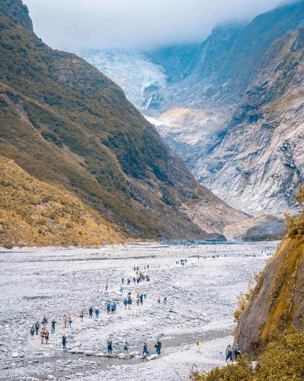 Wide rocky glacier valley with groups of hikers walking toward a glacier surrounded by steep mountains.