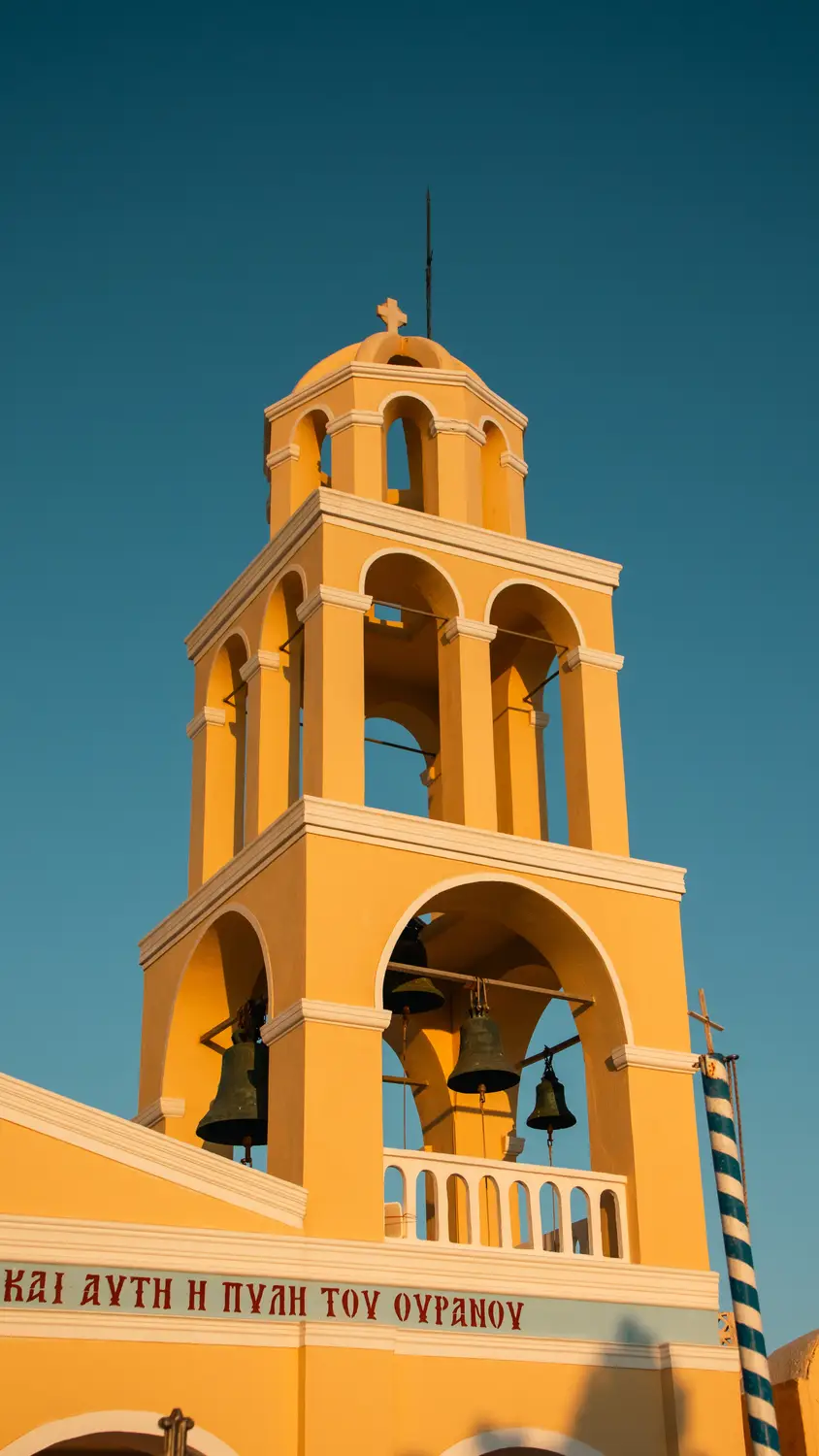 A yellow bell tower of a Greek Orthodox church with several bells, set against a clear blue sky.