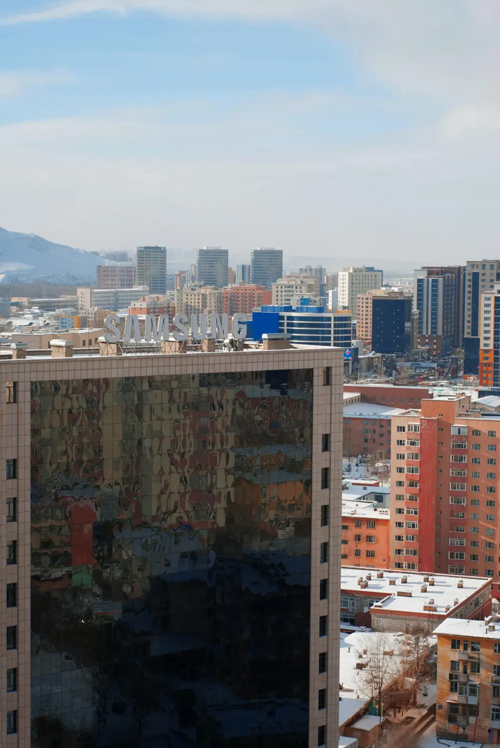 Aerial view of Ulaanbaatar city skyline in winter with snow-dusted rooftops, colorful apartment blocks, and a glass commercial building with Samsung signage, Mongolia
