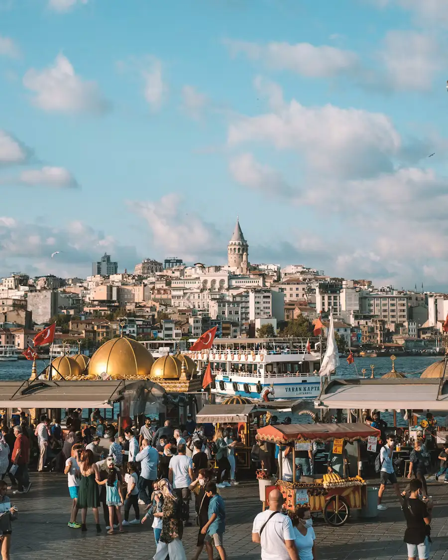 An aerial view of a crowded outdoor market or square in Istanbul, with the Galata Tower and city skyline in the background.