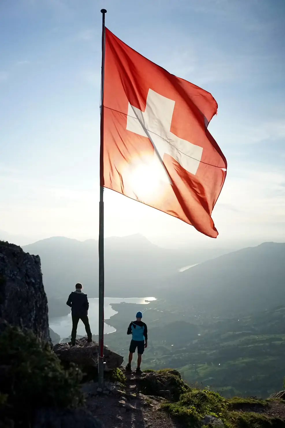 Hikers standing beside a Swiss flag on a mountain summit at sunrise