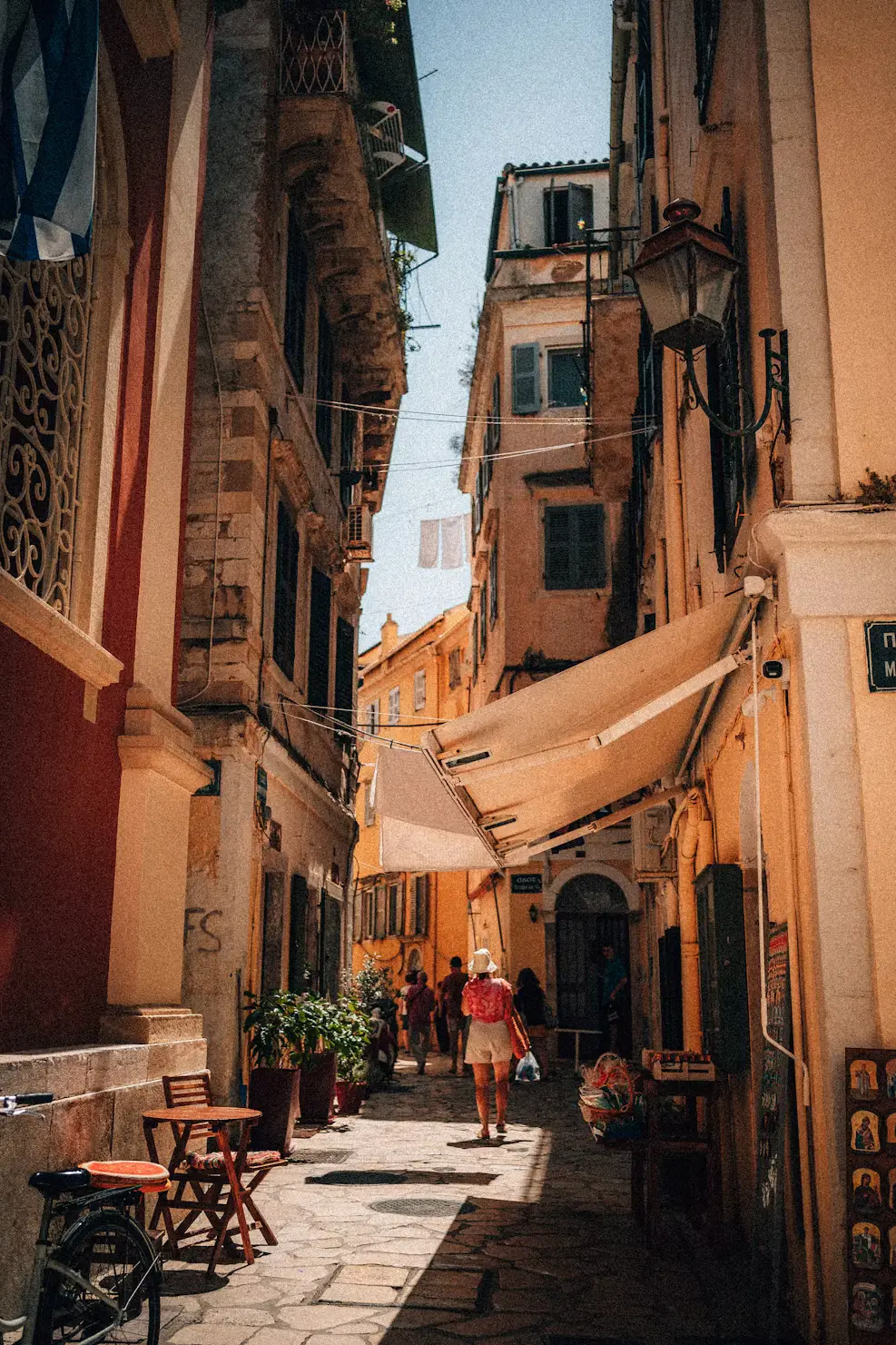 A narrow stone alleyway between old buildings, with outdoor café tables and a person walking past shop entrances.
