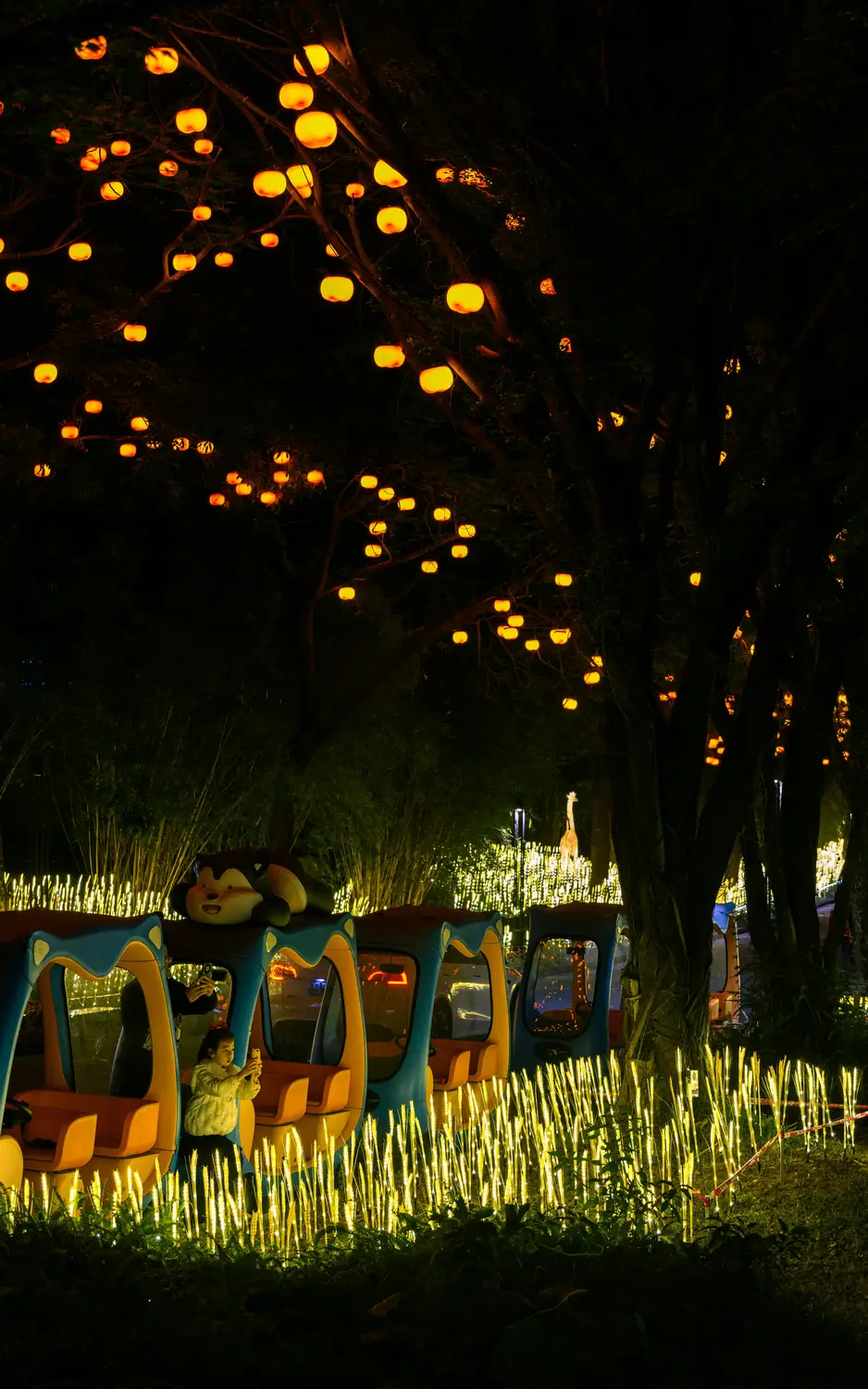 Children's mini train ride through illuminated lantern festival at night with glowing orange lights on trees