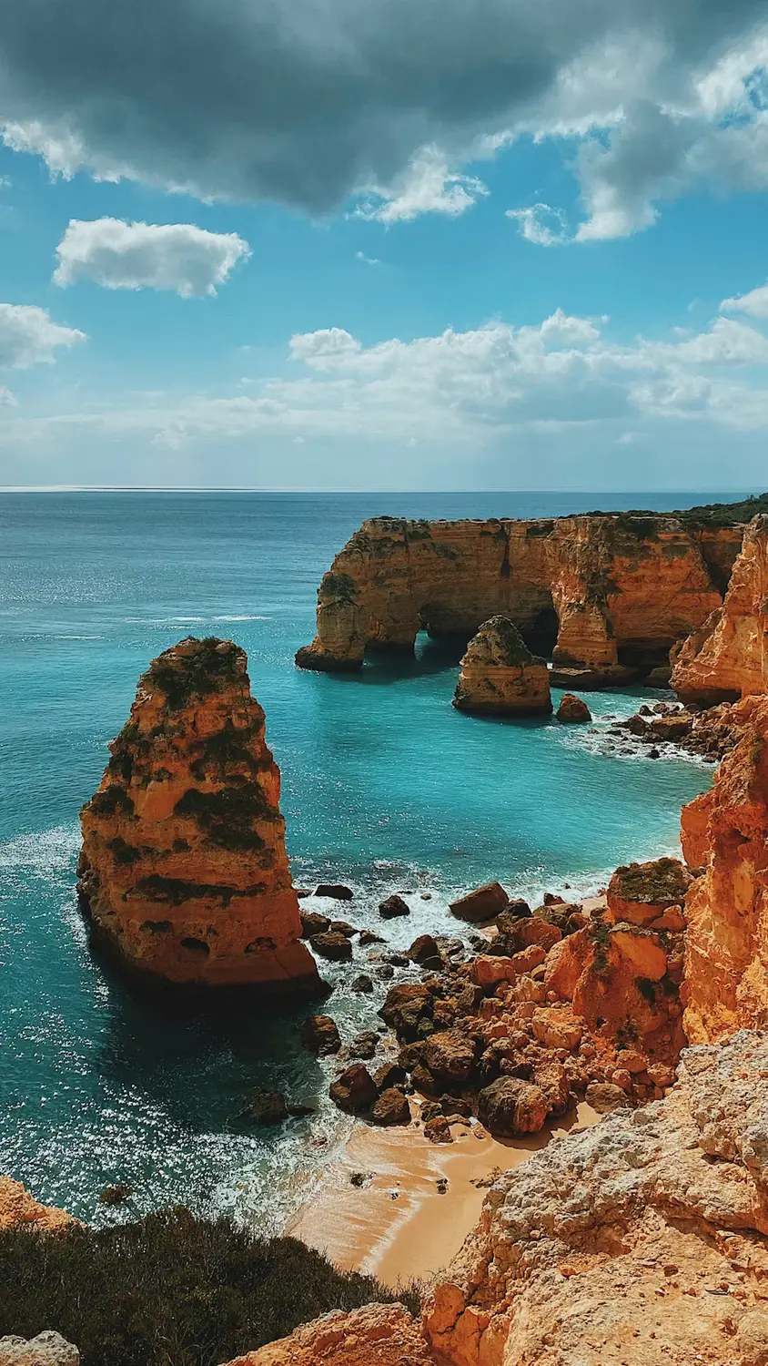 Turquoise water surrounds towering golden sea cliffs and rock formations above a small sandy beach under dramatic clouds.