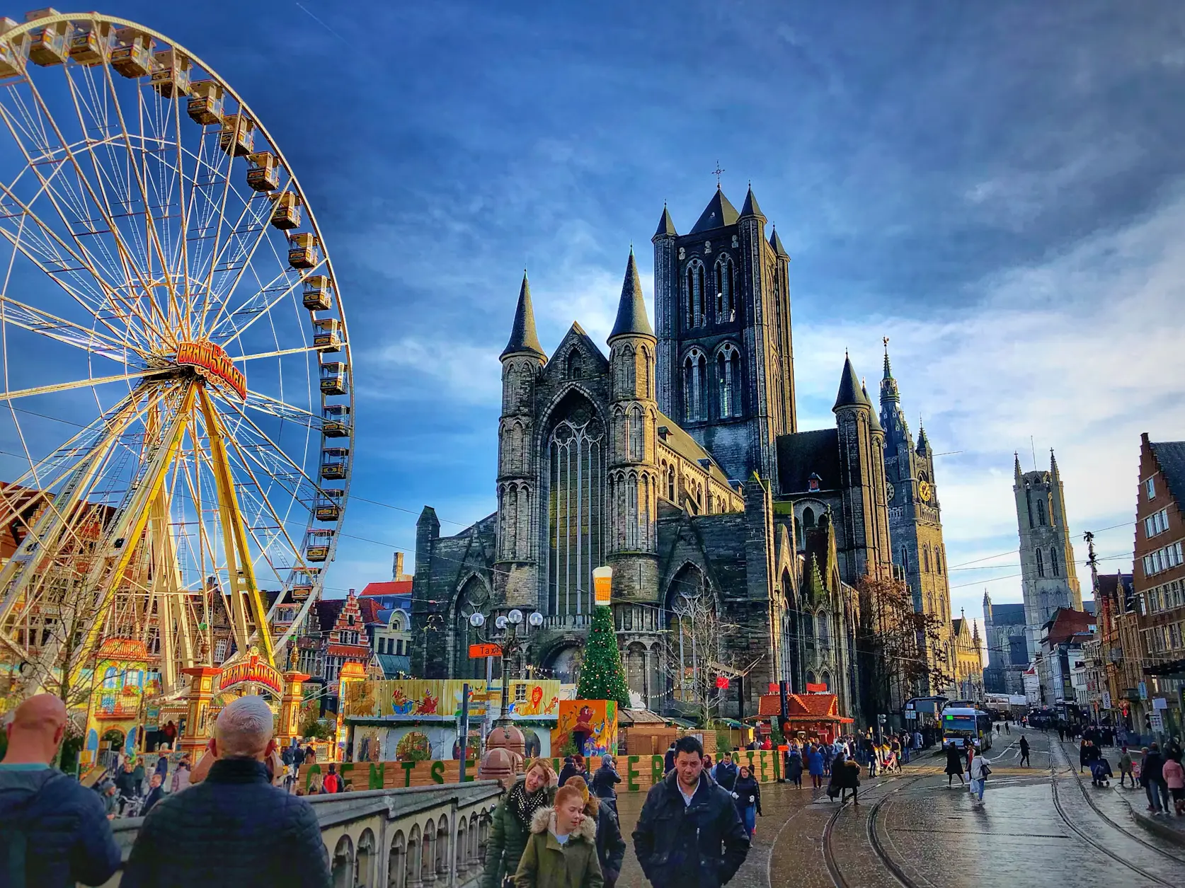 A Christmas ferris wheel beside the Gothic towers of Saint Nicholas' Church in Ghent, with holiday market stalls and pedestrians below.